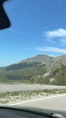 The video shows a car driving on a winding road through a mountainous landscape. The sky is blue with some clouds. There are concrete barriers on the side of the road, and vegetation on the mountains. Two cars pass the camera car, one gray and one red. Someone is talking in the car, asking about a tractor ride.