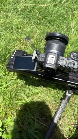 The video begins with a close-up shot of a camera mounted on a slider, positioned on a grassy area. The camera is a Canon, and the slider is branded "TOPPING". The screen of the camera shows the word "PLAY". The scene then transitions to a wider shot of the same camera setup, with sunlight and shadows visible on the grass. Subsequently, the video focuses on the exterior of an old stone building, featuring a wooden door with ornate handles and an arched entrance. The building has rough-hewn stone walls and a single, dark-colored window above the entrance. The camera pans upward, revealing more of the building's facade, including its architectural details and the clear sky above. The video concludes with a shot of the building's facade, emphasizing its weathered stone construction and arched doorways.