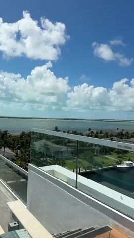 The video begins with a shot of a bright blue sky with scattered white clouds, looking out over a calm body of turquoise water. In the foreground, a modern balcony railing made of glass and white metal is visible. As the camera pans and zooms slightly, the view expands to reveal a resort-like setting with low-rise buildings and lush green palm trees. The water appears to be a channel or bay, with a distant shoreline or island visible. The camera then shifts to focus on a circular, built-in seating area with cushioned seats and small white tables on a wooden deck. Another similar seating area is visible further away. The overall impression is a luxurious outdoor space with a beautiful ocean view.