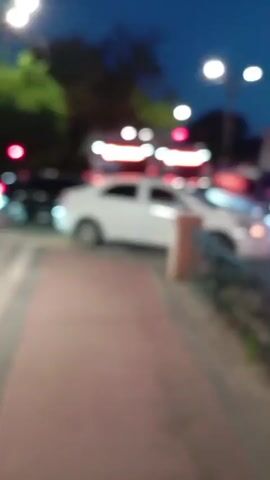 The camera, from the perspective of a sidewalk observer, captures a scene at dusk. Several cars, including a white sedan prominently in the foreground, navigate an intersection. A red bus is visible in the background, further congesting the traffic flow. Streetlights illuminate the scene and cast shadows from nearby trees. An older building with a white facade is visible further down the street.