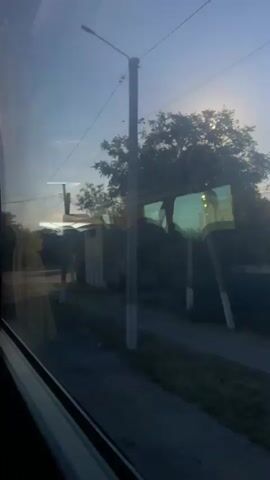 This video captures a view through a window, likely from a vehicle, looking out at a sunlit street scene. The sun is high in the sky, creating a bright glare that obscures some details. On the left, the edge of the window frame and a curtain are visible. Outside, there is a road, a sidewalk, some buildings, trees, and utility poles. The overall impression is of a daytime view in a moderately built-up area, with the sunlight being a prominent feature.