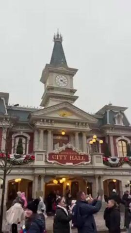 The video shows a busy street scene at Disneyland, presumably the Main Street, U.S.A. area. The architecture is turn-of-the-century American style with a prominent clock tower on the "City Hall" building. The area is decorated for Christmas with wreaths and garlands. Many people are present, bundled in jackets and hats, some with umbrellas. The sky is overcast and there appears to be artificial snow falling, creating a festive atmosphere. People are walking and taking pictures, immersing themselves in the Disney experience.