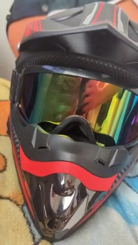 The video shows a close-up view of a black and red dirt bike helmet with rainbow-colored goggles. The helmet is resting on a patterned fabric surface, possibly a blanket or towel. The goggles reflect the image of a person holding a smartphone, presumably taking the video. The lighting is bright, and the focus is on the helmet and goggles, highlighting the reflective quality of the lenses. The background is slightly blurred but appears to be an indoor setting.