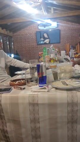 The camera pans from left to right across a table littered with drinks, food containers, plates, and other items. Two men sit at the table, each looking at their respective phones. The man on the left wears a white sweatshirt, while the man on the right is wearing a red t-shirt. On the wall behind them, a TV is playing a video of a woman singing. The room has a rustic feel, with exposed brick walls and a wooden ceiling. A wagon-wheel chandelier hangs above the table. Music plays in the background.