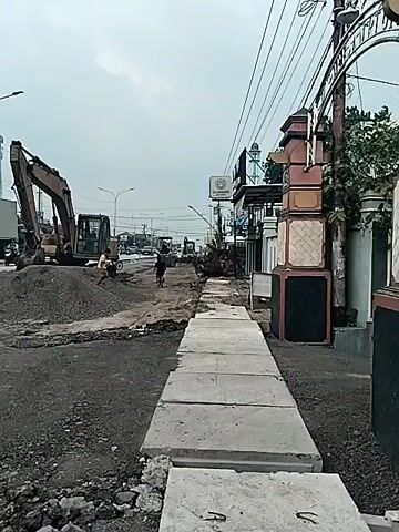 The video shows a street scene in what appears to be a town or city. An excavator is parked on a pile of dirt and gravel in the middle of what looks like road construction. A sidewalk made of rectangular concrete slabs runs along the right side of the frame, leading towards the street. Motorbikes and cars are driving on the road in the background. There is a partially visible gate on the right side of the frame, and power lines run overhead. The sky is overcast, and the general atmosphere suggests ongoing construction or renovation work in the area.