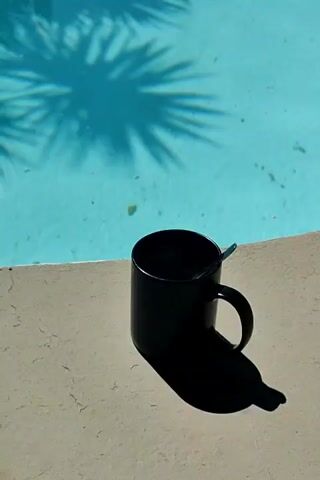 A black mug sits on the edge of a swimming pool. The mug is partially filled with a dark liquid, and a spoon rests inside. Palm tree shadows are cast on the surface of the turquoise water. The sun is shining brightly, creating a sharp shadow of the mug on the pool deck.