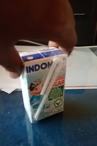A person's hand holds a small white carton of milk, turning it to reveal different sides. The carton has a cartoon cow illustration and text indicating it is "Indomilk Kids Full Cream" and "Tanpa Penambahan Gula." The carton appears to be wrapped in clear plastic. The background shows a blue surface and a white paper with some writing on it.