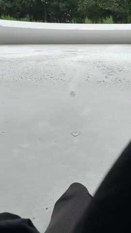 The video shows a close-up view of water droplets on a white surface. The surface appears to be some kind of flat, textured material, possibly a roof or a deck. In the background, there are green trees and foliage, suggesting an outdoor environment. The camera is positioned low, looking across the surface, and the bottom right corner of the frame is blurred by what appears to be a person's leg and clothing. The water droplets are small and irregularly shaped, with some appearing to be in the process of forming or evaporating. There is a sense of stillness and quiet in the scene.