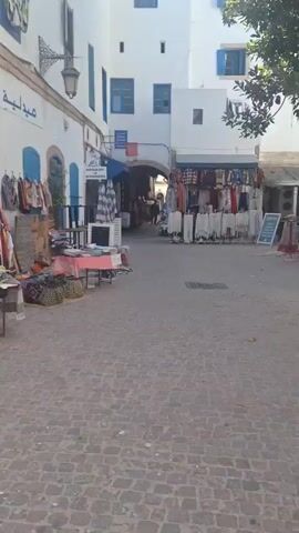 This is a street scene, possibly in a market or souk. The ground is paved with cobblestones. On the left, a white building has blue doors and shutters, and a sign with Arabic script is visible. A display of clothing, possibly garments and scarves, hangs on a rack in front of the building. Bags and other textiles are laid out on the ground. In the center, more items are displayed on tables, including folded fabrics and what appears to be a cooler or an air conditioning unit. Further down the street, an arched passageway leads to more shops or stalls, with people visible in the distance. Another building on the right also features stalls with clothing and other goods. The overall impression is of a bustling marketplace in a town with Mediterranean or North African architectural influences.