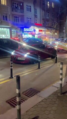The video shows a street scene at night. Several cars are parked or slowly moving. A red car, possibly a sports car, is parked closest to the camera, with its headlights on. The license plate reads "CB 7926 KM." There are a few people visible on the sidewalk. One person is wearing a light-colored jacket and pants, and they are holding a drink can. Several black bollards with white reflective tape are in the foreground, defining the edge of the sidewalk. The street has yellow lines painted on it. Buildings are visible in the background, illuminated by streetlights and neon signs.