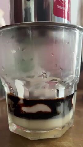 A close-up shot shows a glass of iced coffee with condensed milk at the bottom and dark coffee being dripped from a metal filter on top. The glass has condensation on the outside. A red label with text is visible on the coffee filter. The background is blurred.