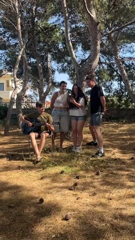 The scene shows four young adults gathered in an outdoor area with tall, bare trees, pine cones on the ground, and a building visible in the background. One person is seated on a chair, playing an acoustic guitar. Three individuals stand nearby, one of whom is holding and looking at a cell phone. The others are also watching and smiling. They appear to be enjoying the music and the moment. The day is sunny, with clear skies and dappled sunlight filtering through the trees.