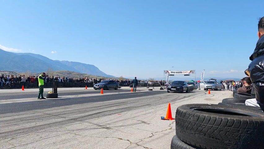 The video shows a drag race between a gray BMW coupe and a black BMW station wagon on an asphalt track lined with orange traffic cones and tire barriers. A large crowd of spectators watches from behind the tire barriers, with a line of mountains visible in the background under a clear blue sky. The vehicles accelerate rapidly, leaving tire marks and some smoke. A race official in a green vest stands near the starting line. There is a "DRAG RACING" banner at the finish line.