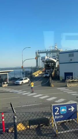 A ferry terminal scene captured from a slightly elevated perspective. In the foreground, a chain-link fence with yellow and red bollards is visible. Beyond the fence, a paved area with pedestrian crosswalks is seen. A person wearing a high-visibility vest and holding a red signaling wand stands in the middle of the road, directing traffic. Several cars are lined up on the roadway, approaching a large white ferry. The ferry is docked at a ramp, with vehicles visible driving onto its deck through an open entryway. The sky above is clear and blue. A sign with the number "2" and a ferry icon is partially visible to the right, indicating berth number two. The overall impression is one of organized embarkation at a ferry terminal under clear, sunny conditions.