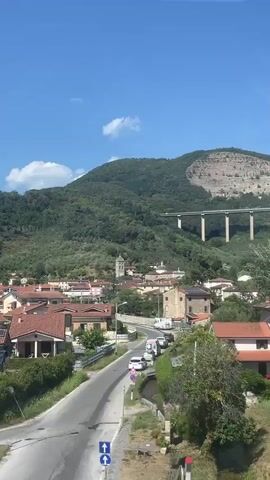 A scenic view of a hillside town in Italy under a clear blue sky. The town is nestled amongst lush green trees and rolling hills. In the background, a modern highway bridge with concrete pillars stretches across the landscape, traversing a mountainous area. The buildings in the town feature red-tiled roofs and cream-colored facades. The foreground shows a grassy field with some trees and what appears to be a small stream or ditch. The overall impression is one of a peaceful rural setting with a touch of modern infrastructure. There are no people visible in the video.