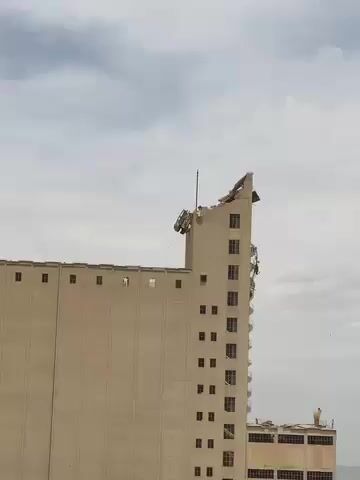 The video opens with a view of a tall, light-colored concrete building, possibly a grain silo or an industrial structure, against an overcast sky. The very top of the building appears damaged or partially demolished, with some structural elements visibly broken or missing. The camera then pans down briefly to show a lush green bush or tree with long, narrow leaves, before panning back up to the building. Another, shorter building is partially visible in the background to the right. A tall, slender evergreen tree stands on the left side of the frame as the camera focuses again on the damaged building. The scene takes place during the daytime in what appears to be an urban or industrial area in Panjwai, Afghanistan.