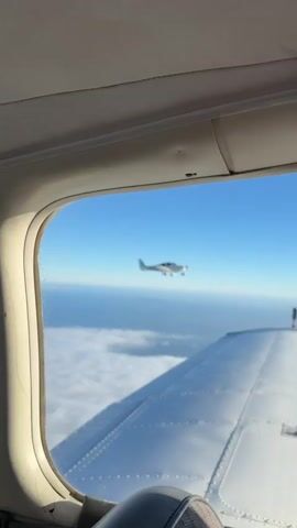 This is a video of a small white airplane flying in the sky. The airplane is seen from the perspective of another airplane, looking out of a window. In the foreground, a portion of a wing from the viewpoint airplane is visible. In the background, the sky is blue, and there are clouds below. The airplane in flight has the registration number N219FI on its tail. The video shows the airplane from different angles as it flies alongside.