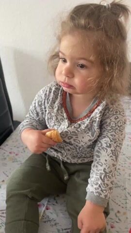 A close-up shot shows a toddler sitting on a patterned surface, possibly a bed. She is holding a piece of croissant in her right hand and looking towards the camera with a slightly serious expression. The girl has curly brown hair, styled in a small top ponytail, and is wearing a gray patterned long-sleeved shirt with a red trim around the collar, and green pants. The background is a plain white wall. The shot is well-lit and stable.