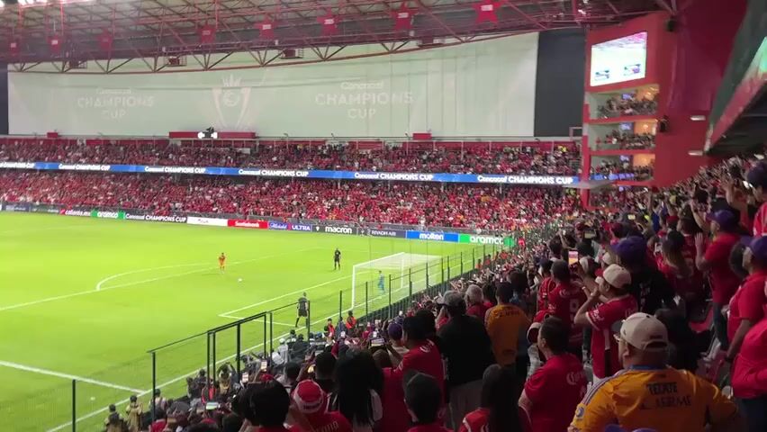 The video shows a soccer match in a stadium filled with fans. The stadium is mostly red, with a large banner that reads "CONCACAF CHAMPIONS CUP." The field is green and the players are in uniform. The fans are wearing red and other colors, and many of them are holding up their phones to record the game. The atmosphere appears lively and energetic.