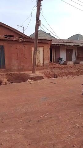 The video shows a street scene in what appears to be a rural or developing area. The road is made of red dirt and is uneven. There are simple, weathered buildings lining the road, some with corrugated metal roofs. A utility pole with wires is visible in the foreground. Several motorcycles, each carrying one or two people, are driving down the road. The sky is overcast with a hint of sunset glow. The overall impression is that of a typical day in a simple, possibly impoverished, community.