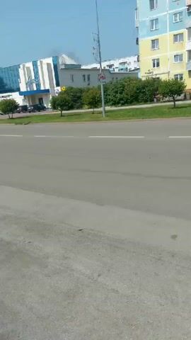 The video shows a street view in front of buildings. On the left, there is a light-colored building with a blue stripe and a white section. Next to it are several trees and a yellow apartment building. In the center, a modern building with a glass facade and white accents is visible. Several cars are parked in front of this building. There are also trees and a street sign. The road in the foreground is asphalt with white lane markings. The sky is clear and blue.