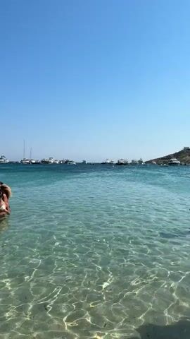 The video shows a clear, turquoise ocean with sunlight reflecting off the sandy bottom. Several yachts are anchored in the distance, and a small group of people are visible swimming or wading in the shallow water near the shore. The sky is clear and blue.