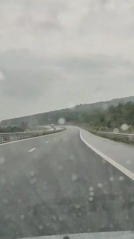 A POV shot from inside a vehicle driving on a wet, multi-lane highway. Rain is visible on the windshield, appearing as blurred circles. The sky is overcast with grey clouds. To the left, there is a metal guardrail and trees lining the road. In the distance, there are hills covered in green foliage. Several vehicles are visible on the highway, including trucks and cars. The road surface is dark asphalt with white lane markings. The overall atmosphere is gloomy and damp due to the rain.
