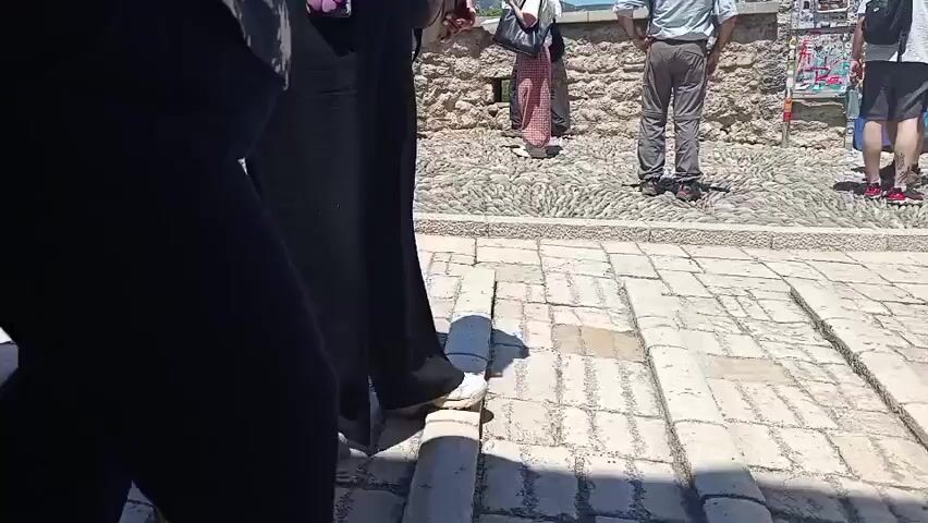 The video shows a close-up view of people walking on a cobblestone street. The focus is on their legs and feet, suggesting a perspective from someone walking behind them or filming from a low angle. The subjects are dressed in casual summer clothing. One person wears black pants and white sneakers, another is in patterned trousers and light-colored shoes, and a third person is seen in a dark skirt and what appear to be sandals. In the background, other people are visible, some in jeans and t-shirts, and a glimpse of a stone wall and some posters can be seen. The ground is paved with irregular-shaped stones, with some sections of larger, flatter stone blocks. The lighting suggests it is daytime, likely with bright sunlight, casting distinct shadows on the ground. The overall atmosphere appears to be that of a tourist area or a town square, with people casually strolling. The cobbled street and stone architecture in the background are indicative of a historic European setting, consistent with the provided location context of Mostar, Bosnia and Herzegovina. There is no visible text in the video.