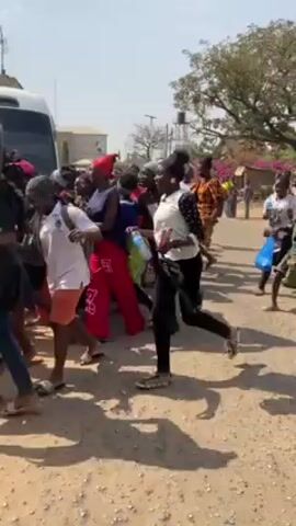 The video shows a crowd of people, primarily women, running toward or away from a white van or bus on a dirt road. Some are carrying items like bags. The crowd appears energetic and chaotic. In the background, there are buildings and trees under a bright sky. The road is unpaved and light brown in color. The camera is moving along with the crowd.