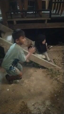 The video appears to be taken at night, possibly in a rural or semi-rural setting in Keude, Indonesia. The scene is dimly lit, with artificial light sources casting shadows and highlighting certain areas.

The primary subject is a young boy, who is the focus of the initial frames. He is wearing a light-colored t-shirt and shorts, and appears to be in motion, possibly playing or running. He is wearing sandals. Another person is partially visible in the background, also appearing to be moving.

Later in the video, a motorcycle becomes a prominent object. It is red and black, and its design suggests it's a relatively modern model. A person, likely an adult, is seen interacting with the motorcycle, possibly preparing to ride it or having just arrived. This person is wearing a light-colored shirt and pants and is also wearing sandals.

The setting seems to be outdoors, with a mix of dirt and concrete ground. There are structures in the background that suggest a shop or a place of business. One of these structures has a sign with text that reads "BAKSO SUPRA". This indicates the presence of a food stall, likely selling bakso (meatball soup), a popular Indonesian dish. The sign is brightly lit, contrasting with the surrounding darkness.

The overall mood is casual and everyday. The activities appear to be routine, such as children playing and someone preparing to use a motorcycle. The time of day being night contributes to the atmosphere, with the artificial lights creating a warm glow in some areas. There are no obvious signs of distress or urgency; the activities seem relaxed.