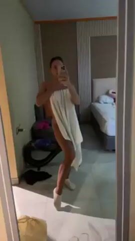 A woman standing in a bathroom, mostly covered by a white towel, takes a mirror selfie with her cell phone. She is wearing white socks and her hair is pulled back. Behind her, there is a bed, a chair with clothes on it, and a bag on the floor. The bathroom floor is tiled. The room appears to be a hotel room.
