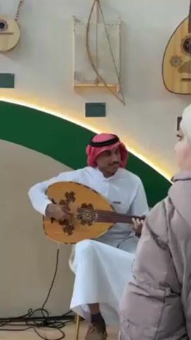 A man wearing traditional Arab clothing and a red and white headdress is seated and playing an Oud, a pear-shaped stringed instrument. He is surrounded by other Oud instruments displayed on the wall behind him, which is adorned with a green wavy shape and illuminated by a light strip. He is smiling as he plays. Another person, possibly a woman wearing a gray jacket, is standing near him. The setting appears to be a museum or exhibit showcasing traditional instruments.