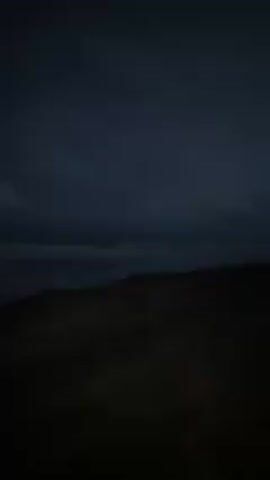 The video shows a dark beach scene, likely at dusk or night, with a view of the ocean and a cloudy sky. The camera pans slightly, showing the horizon and distant lights. The beach is dark and indistinct, with the focus primarily on the sky and the breaking waves.