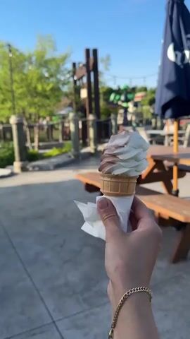 This still image captures a person's hand holding a soft-serve ice cream cone in an outdoor setting, likely in Chelsea, Canada. The ice cream is a classic vanilla and chocolate swirl, served in a waffle cone and wrapped at the base with a white napkin. A golden chain bracelet adorns the wrist of the hand holding the cone.

The background reveals a sunny outdoor patio or recreational area. Several wooden picnic tables are visible, along with a large blue umbrella providing shade. Beyond this area, green trees, rustic wooden posts, and other structures can be seen, suggesting a park, a mini-golf course, or an outdoor eatery. The ground is paved with concrete. The clear, bright blue sky indicates good weather, likely late afternoon or early evening given the lighting. There is no visible text in the image.