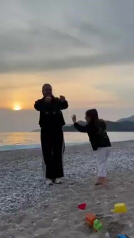 On a pebble-strewn beach at sunset, a woman and a young girl are interacting. The woman, wearing a black hooded jacket and black pants with a white stripe down the side, has her hair pulled back. The girl, dressed in a black jacket and white pants, reaches out to the woman, pulling at her arm. The woman takes the girl's hand, then places her other hand on the girl's waist, holding her in a playful embrace. The sun sets over the ocean in the background, casting a warm glow on the scene. Scattered on the beach are various plastic toys, including a red heart, an orange cube, and a green and orange toy truck. The sky is cloudy, and the ocean is calm.