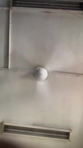 The video shows a white ceiling fan spinning rapidly in the center of the frame. Above and below the fan are two rectangular ventilation grilles, also white. The ceiling is painted a light color. Pipes run along the ceiling and wall in a grid-like pattern. The fan's blades are blurred due to their speed. The overall scene appears to be an indoor environment, possibly an office or classroom.