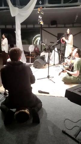 A group of people are gathered indoors, possibly at an event or performance. In the foreground, a person wearing a black hoodie and dark pants sits on the floor playing a drum. Behind them, a woman stands and sings into a microphone, holding a tambourine. Two other individuals are seated, one playing a guitar and the other appearing to be singing or playing a stringed instrument. Other instruments and equipment, including microphones and speakers, are visible. The background features draped white fabric and some decorative lighting. The setting appears to be a covered area, possibly a tent or open-sided building.