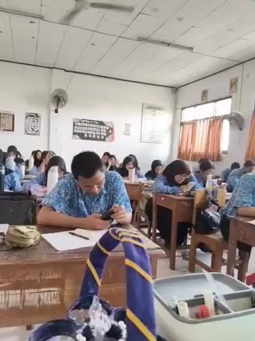 This is a video taken inside a classroom. The camera is focused on a male student in the foreground, wearing a blue patterned shirt, who is looking down at his phone while sitting at his desk. There are other students, mostly female, seated at desks in rows behind him. Many of them appear to be wearing hijabs and school uniforms. The classroom has white walls, a ceiling fan, and windows with orange curtains. There are posters and charts on the walls. On the desk in front of the male student, there is a notebook, a pen, a bottle of water, and other items. The overall impression is of a typical school environment during a lesson.