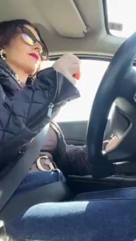 A woman is driving a car. She has dark hair and is wearing sunglasses, a black quilted jacket, blue jeans, a large G buckle belt, and red lipstick. The woman exhales a cloud of white vapor while driving.  The sun is shining and the sky is bright. The video appears to be taken from the passenger seat.