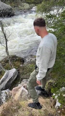 A single man wearing a grey sweatshirt, light olive green pants, and black shoes is walking down a rocky hill towards a white-water river. He navigates the rocks with care, occasionally using a tree for support. The river is turbulent and loud, creating a dramatic contrast to the man's calm movements. The environment consists of trees, rocks, and some dry grass and moss. The shot is stable and focuses primarily on the man and the river.