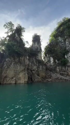 This media file captures a scenic view from a boat on a body of water, likely a lake, with a backdrop of dramatic karst formations. The water is a vibrant turquoise, reflecting the sky and the surrounding cliffs. The cliffs themselves are rugged and steep, covered in lush green vegetation and trees, particularly on their upper reaches. The sun is setting or rising, casting a warm glow and lens flares on the left side of the frame, highlighting the textures of the rock faces. The sky is partly cloudy, with the clouds creating interesting patterns and adding to the atmospheric quality of the scene.

The prominent geological features are the tall, jagged limestone peaks that rise directly from the water, some with precarious-looking trees clinging to their summits. The scale of these formations suggests a vast natural landscape. The activity taking place is implied to be a boat journey or a leisurely cruise through this breathtaking natural environment.

The time of day is either early morning or late afternoon, indicated by the low angle of the sun and the quality of light. The weather appears pleasant, with a mix of sun and clouds. There is no visible text in the media file. The overall emotion evoked is one of awe and tranquility, emphasizing the beauty and grandeur of nature. The location cues, such as the distinctive karst topography and lush vegetation, align with the provided context of Khao Phang, Thailand, known for its stunning limestone cliffs and emerald waters.