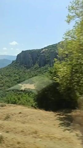 A video shows a mountainous landscape with lush green trees covering the slopes. The sky is clear blue with a few white clouds. The camera appears to be moving, possibly from a vehicle, as the foreground shows a grassy, dry slope that blurs slightly. In the distance, layers of mountains fade into the horizon. The prominent feature is a large rock formation or cliff face on the side of the mountain. The video is brief and appears to be a scenic travel shot.