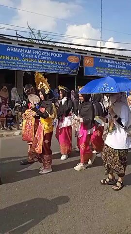 A group of people parade down a street. On the left side of the frame, several girls wear black hijabs and colorful outfits including gold headpieces and pink and gold skirts.  A woman in a white t-shirt and patterned skirt holds a blue umbrella over them.  To the right, a group of boys in white shirts and patterned black skirts walk in a line. They wear black peci hats.  A crowd of people watches the parade from the side of the road. Banners advertising ARMAHAM FROZEN FOOD and ARMAHAM PLASTIC SHOP hang above the parade route. The sky is blue with scattered clouds.