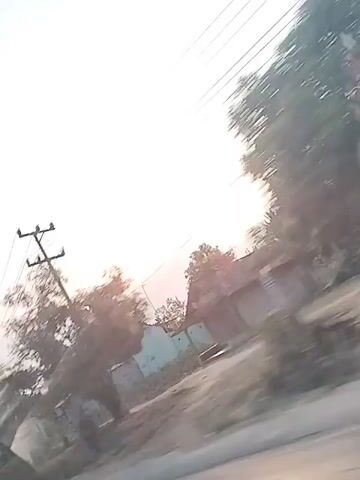 The video shows a view from a moving vehicle looking upwards towards the sky. Overhead power lines stretch across the frame, supported by utility poles. The sun is visible, low in the sky, casting a soft glow through a hazy atmosphere. Silhouetted trees and buildings are visible along the bottom of the frame, suggesting a semi-urban or rural setting. The motion of the vehicle causes a slight blur, particularly in the foliage and structures, but the power lines and sky remain relatively stable. There are no people visible in the video. The overall impression is of a somewhat overcast or dusty day with the sun attempting to break through.