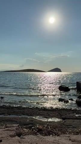 A still shot of a bright sun over the ocean, with a faint shimmering reflection of light on the water. Two small islands are visible in the distance. The foreground shows a rocky shore with some debris and a small puddle of water.
