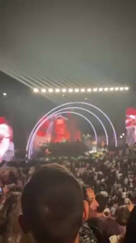 The video captures a view from the back of a large crowd attending a concert. On a brightly lit stage, a band is performing, with the lead singer visible on large screens on either side of the stage. The stage setup includes arched lighting structures. The audience appears dense, and some individuals are looking towards the stage with smiles. The lighting is bright, suggesting the event is either well-lit at night or being held indoors. The crowd seems to be enjoying the concert.