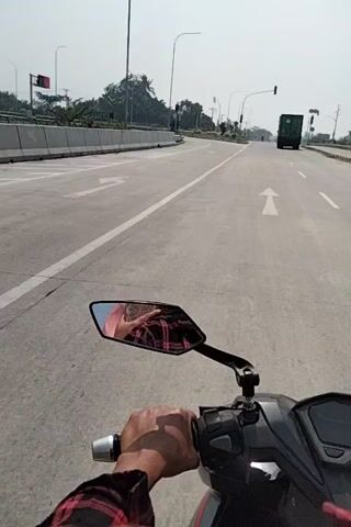 The video is shot from the perspective of someone riding a scooter on a highway. The camera is mounted on the scooter, pointing forward and slightly to the right, capturing the rider's hand on the handlebar and a rearview mirror. The highway appears to be newly constructed, with clear white lane markings and a concrete barrier on the left side. Trees and some infrastructure, possibly lights or traffic signals, are visible in the distance. The rider's reflection in the rearview mirror shows them wearing a plaid shirt. A truck is visible in the far distance on the highway. The sky is overcast, suggesting a cloudy day. The video is shaky, indicating movement.