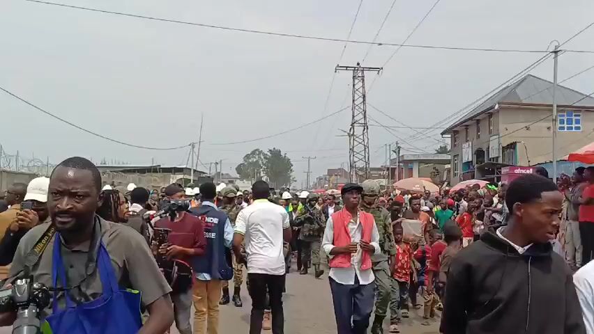 A large crowd of people is walking down a street, some wearing military uniforms and carrying rifles. There are also people wearing hard hats and vests, and some are in civilian clothes. The street is lined with buildings and shops, and there are power lines overhead. The sky is overcast.