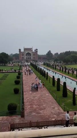 The video shows an aerial view of a well-maintained garden with a central pathway leading to a large, ornate building. The pathway is made of red paving stones and is lined on one side by manicured green lawns with rounded bushes and on the other by a long, narrow reflecting pool. Tall, conical trees are spaced along the edge of the pool. Numerous people are walking along the pathway towards the building, which appears to be a gateway or entrance to a larger structure. The sky is overcast, suggesting a cloudy or hazy day. The overall impression is of a tourist destination with significant foot traffic.