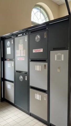 The video shows a bank of gray and black luggage lockers with small white suitcase icons. The lockers are different sizes and have various labels and numbers. The camera pans slowly to the right along the row of lockers, partially revealing another bank of lockers, then the window. The location appears to be indoors with tiled floors and pale walls.
